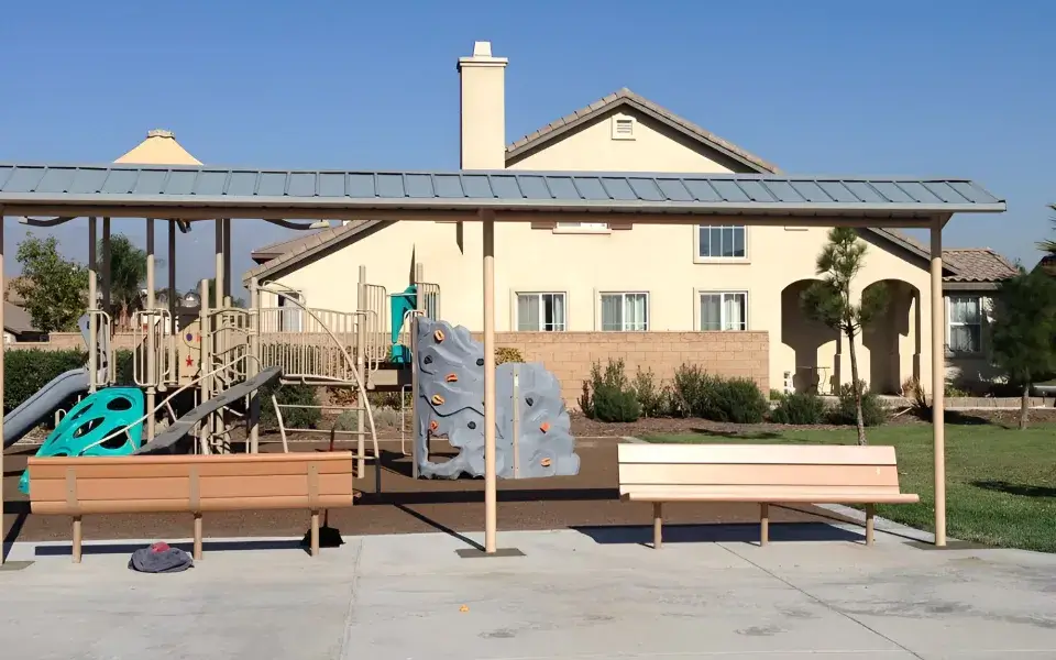 Playground structure with climbing wall and slides, shaded seating area, and residential backdrop in Hemet, California.