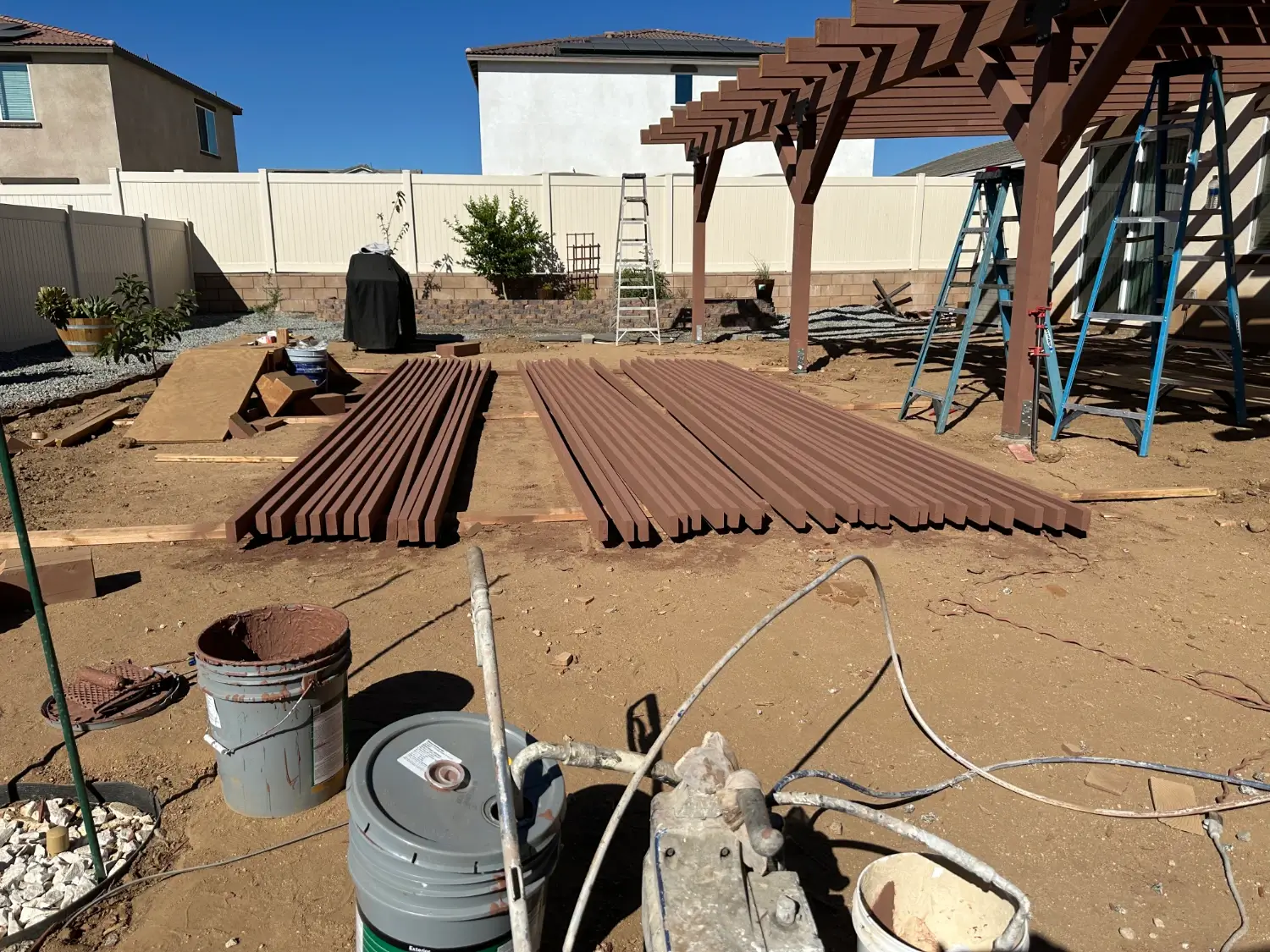 Construction site in Ontario, CA featuring stacked brown lumber for patio cover installation, surrounded by tools and equipment, with residential homes in the background.