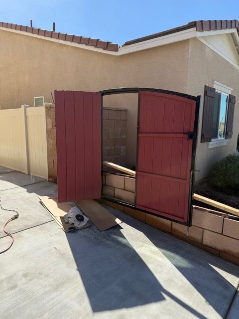 Custom-built red wooden gate with an arched top, partially open, installed beside a residential property, showcasing craftsmanship and tailored design for Riverside, CA conditions.