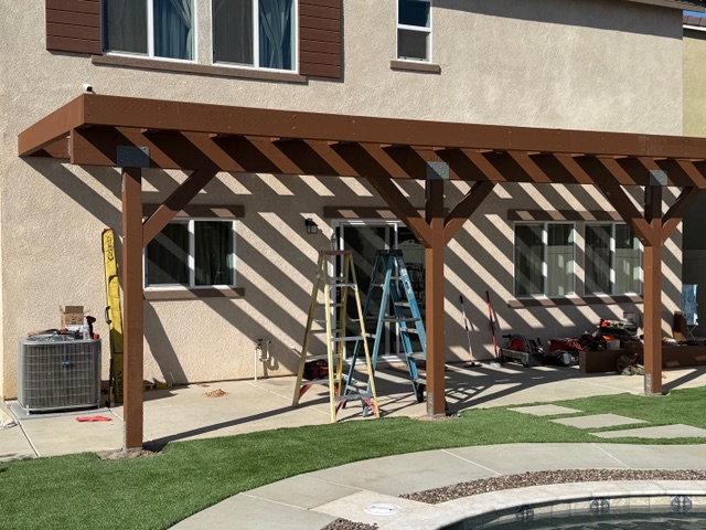 Wood patio cover construction in progress, featuring brown beams, ladders, and tools, adjacent to a residential home in Riverside County.